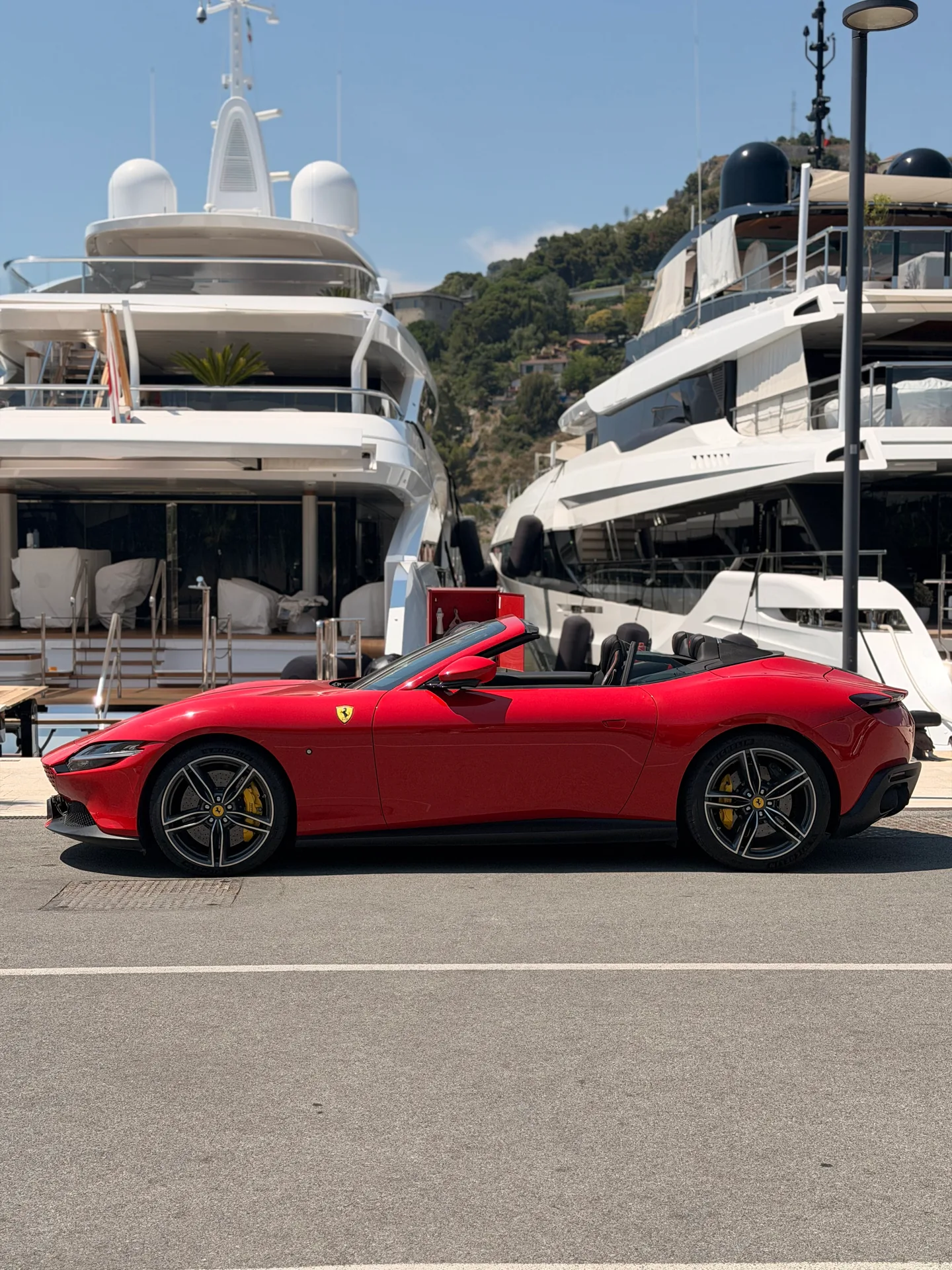 Profilo laterale di una Ferrari Roma Spider rossa a tetto aperto accanto agli yacht.